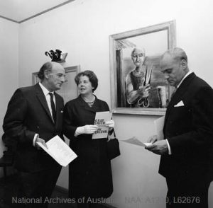 Black and white photograph of Gray Smith, Zara Holt and Henri Souillac standing together at the 1967 preview exhibition in Souillac's Canberra home, Zara Holt holding the exhibition programme, a framed portrait on the wall behind them.