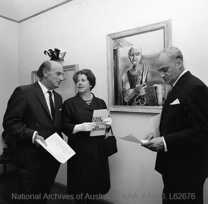 Black and white photograph of Gray Smith, Zara Holt and Henri Souillac standing together at the 1967 preview exhibition in Souillac's Canberra home, Zara Holt holding the exhibition programme, a framed portrait on the wall behind them.