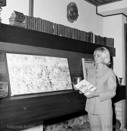 Black and white photograph of an unidentified blonde woman in a pale suit holding the exhibition programme at the 1967 preview in Henri Souillac's Canberra home, standing before a wide painting mounted above a brick fireplace on a timber-panelled wall.