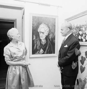 Black and white photograph of a woman in a satin dress, believed to be Lady 'Molly' Huxley, talking with Henri Souillac at the 1967 preview exhibition in his Canberra home, standing either side of Gray Smith's portrait of Lady Huxley.
